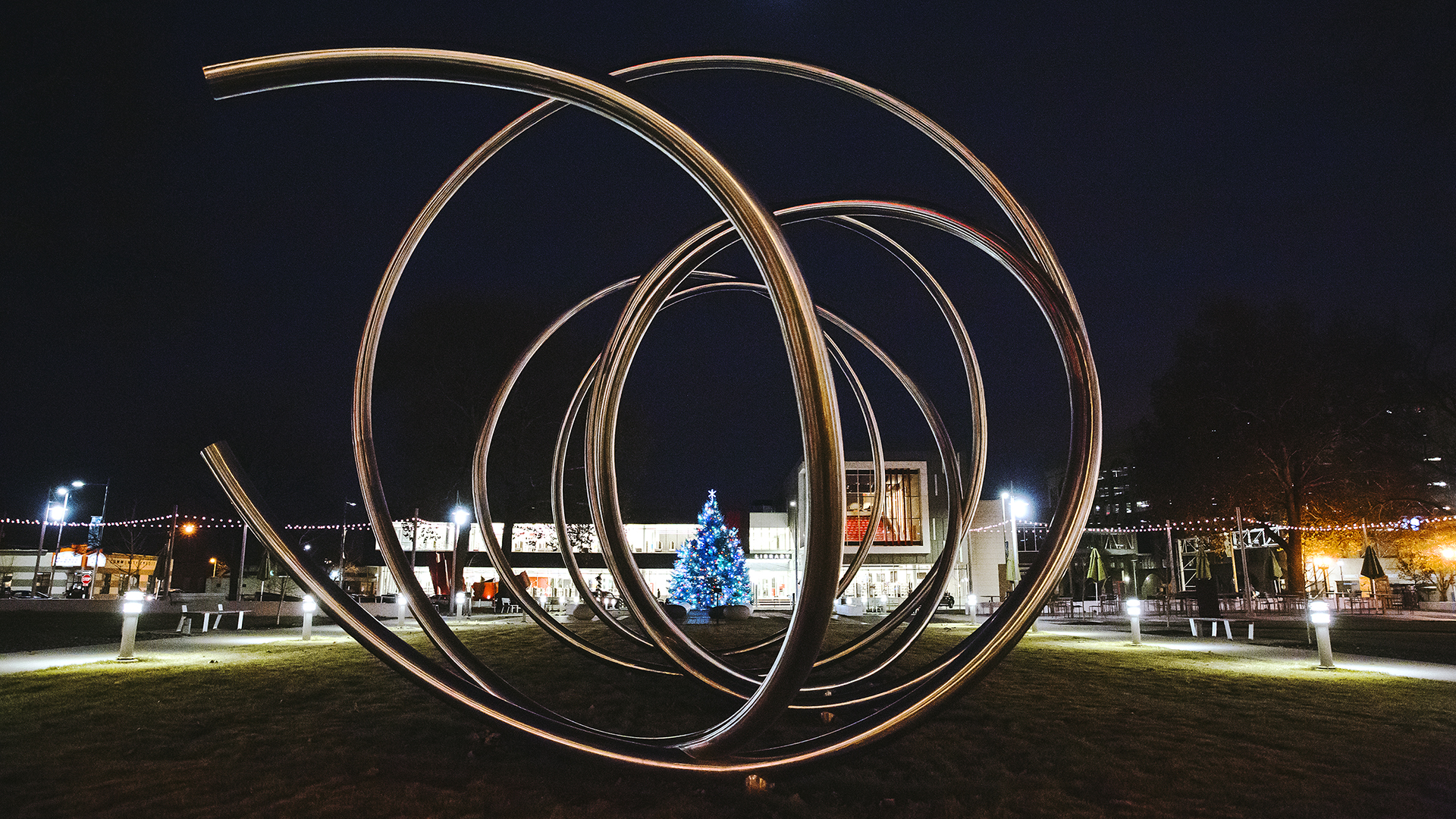 A Christmas tree centered behind a metal sculpture surrounded by Christmas lights at night