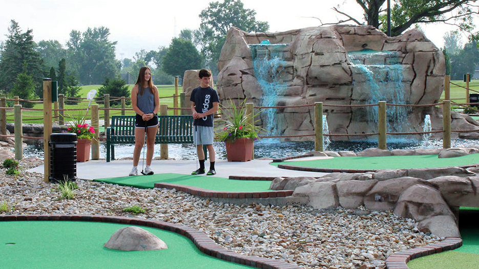 a teenage boy and girl stand with golf clubs while waiting to mini golfing at mini pines in cedar rapids, IA